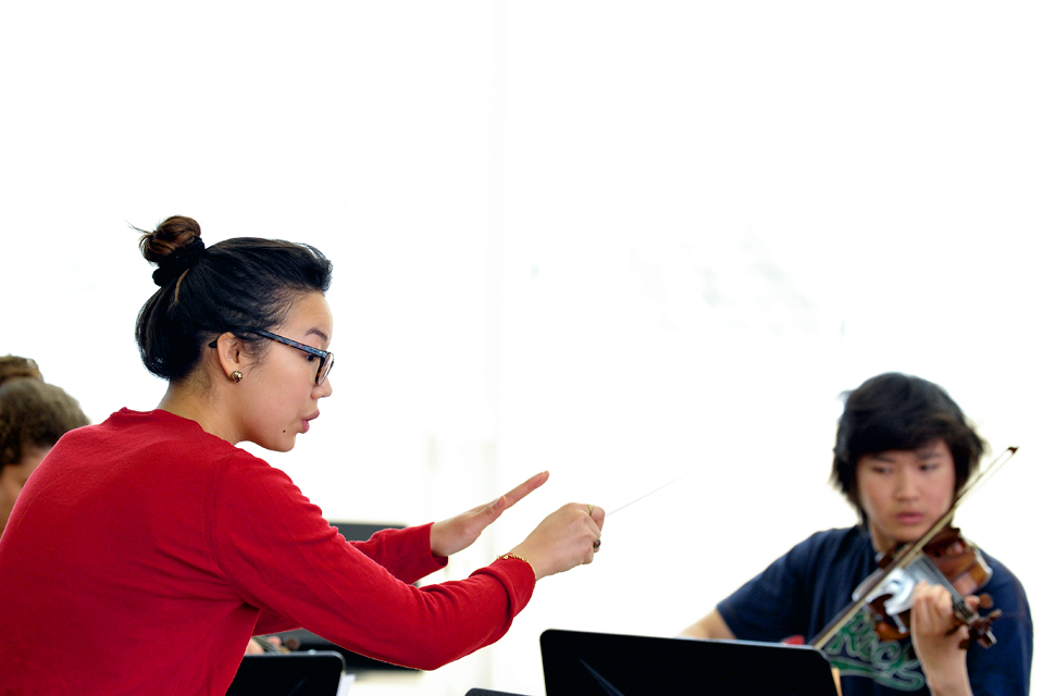 A female student, looking at their music sheets, using a conductor's baton to conduct a group of students performing in a rehearsal.